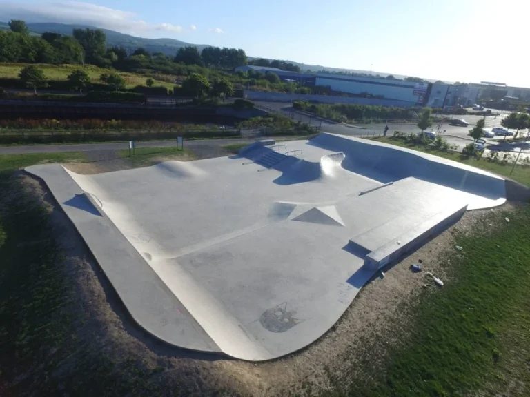 Aerial view of a large outdoor concrete skatepark with ramps, rails, and ledges, surrounded by grass, parking lot, and trees